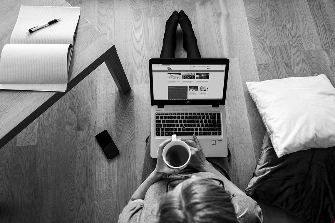 En person sitter på golvet med datorn i knät och en kopp kaffe i händerna.